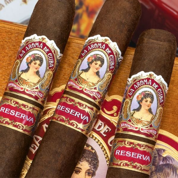 Three La Aroma De Cuba Reserva Belicoso cigars with ornate bands featuring a woman’s portrait and rich San Andrés Oscuro wrappers rest on a wooden cigar box.
