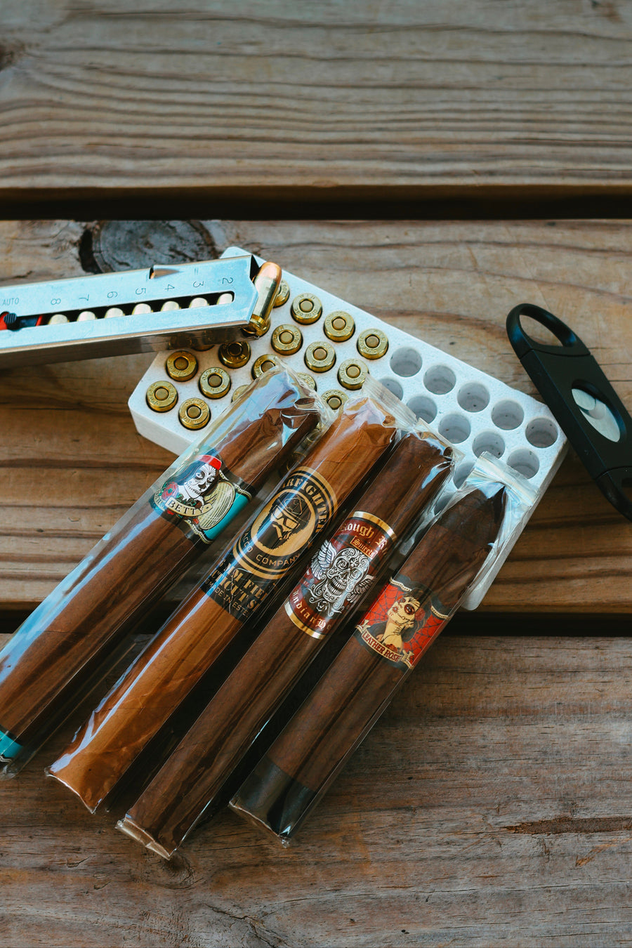 Four cigars in wrappers, a cigar cutter, and a tray of bullets with a revolver speedloader on a wooden surface.