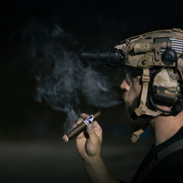 In the dark, someone in tactical gear and night vision goggles enjoys an After Action Cigars TAG x After Action Limited-Edition Cigar (5-Pack), smoke swirling around them.