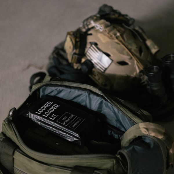 A tactical backpack with a pouch labeled "LOCKED. LOADED. LIT." sits open beside a military helmet, binoculars, and an After Action Cigars TAG x After Action Limited-Edition Cigar (5-Pack) on the ground.