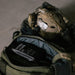 A tactical backpack with a pouch labeled "LOCKED. LOADED. LIT." sits open beside a military helmet, binoculars, and an After Action Cigars TAG x After Action Limited-Edition Cigar (5-Pack) on the ground.