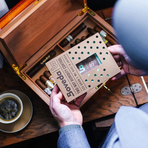 A person holds a Boveda 69% Humidity Control Pack - Single (320g) above an open humidor of cigars, ensuring ideal humidity with Boveda, as a cup of coffee sits nearby.