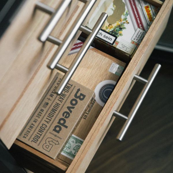 An open wooden drawer displays cigar boxes, a Boveda 69% Humidity Control Pack - Single (320g) by Boveda, and other cigar items—perfect for those who need large humidors and dependable humidity control.