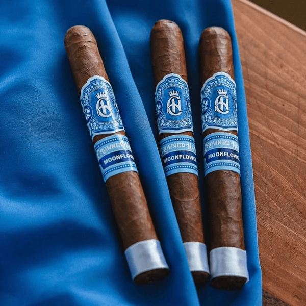 Three Crowned Heads Moonflower Corona Gorda cigars, each adorned with blue and silver labels and wrapped in premium Nicaraguan Corojo leaf, rest on a blue cloth beside a wooden surface.