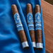 Three Crowned Heads Moonflower Corona Gorda cigars, each adorned with blue and silver labels and wrapped in premium Nicaraguan Corojo leaf, rest on a blue cloth beside a wooden surface.