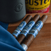 Three blue-labeled cigars, including the distinguished Crowned Heads Moonflower Corona Gorda wrapped in a premium Nicaraguan Corojo wrapper, rest on a wooden surface near a black cup, a blue cloth, and a yellow canister in the background.
