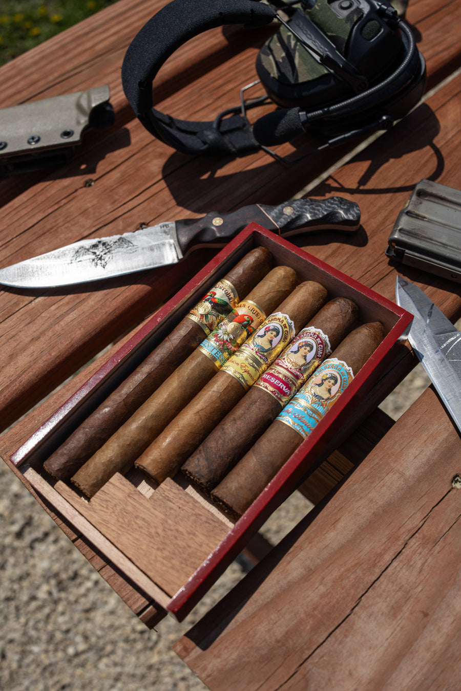 A wooden box with five assorted cigars sits on a picnic table next to a knife, headset, and other outdoor gear.
