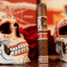 A Deadwood Dominicana Buenas Noches Robusto cigar with a sugar skull band, positioned between two painted skull figurines and a red rose.