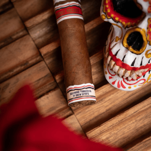 A Deadwood Dominicana Buenas Noches Robusto cigar with a white, red, and blue band placed next to a decorated skull figurine on a wooden surface.