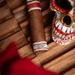 A Deadwood Dominicana Buenas Noches Robusto cigar with a white, red, and blue band placed next to a decorated skull figurine on a wooden surface.