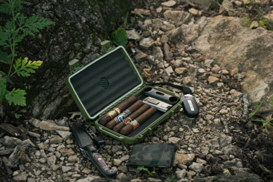 A green cigar case holding cigars and accessories sits open on rocky ground outdoors, next to a folding knife, wallet, and lighter.