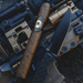 A limited-edition cigar, the Spartan Blades Habano Toro, rests beside a black tactical knife on a tan and black firearm with visible attachments, perfectly capturing the bold character of its namesake.