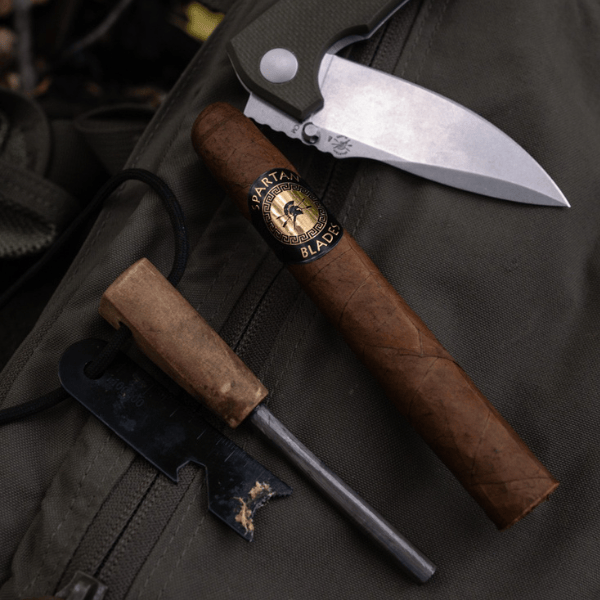 A closed folding knife, a Spartan Blades Habano Toro brown limited-edition cigar, and a fire starter with striker sit atop an olive-green backpack.