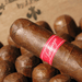 Close-up of a Tatuaje Havana VI Nobles cigar with a red "Tatuaje" label and an Ecuadorian Habano wrapper, surrounded by several other cigars in the background.