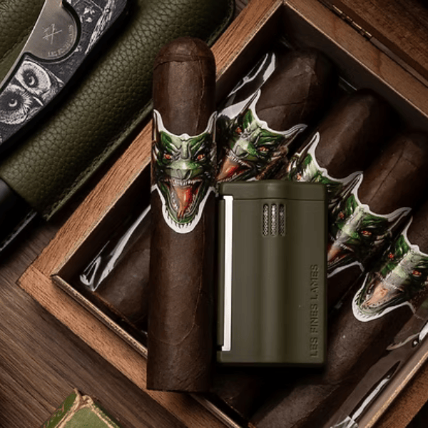 A box of Big Sky Cigar Cryptid Gordo cigars with green dragon face bands featuring a Mexican San Andrés wrapper, accompanied by a cigar cutter and a matte olive lighter, are arranged neatly on a tabletop.